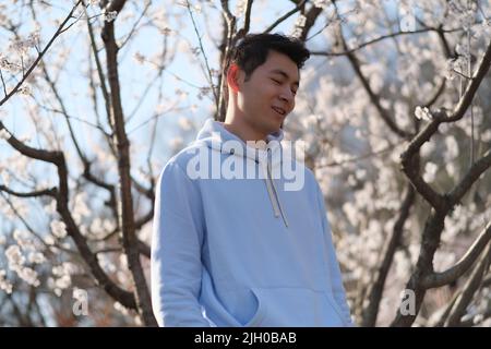 Hübscher junger Mann aus Asien blickt geschlossen und lächelt, genießt das Sonnenlicht unter dem weißen Sakura-Blütenbaum Stockfoto