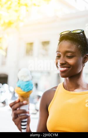 Porträt einer afrikanerin, die dem bunten Eis auf der Hand sieht. Sie trägt Sommerkleiderobe und lächelt Stockfoto