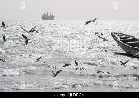 PORTUGIESISCHE STADT, BANJUL, GAMBIA - 10. FEBRUAR 2022 Containerschiff, Möwen und festgemacht lokalen Fischerboot Stockfoto