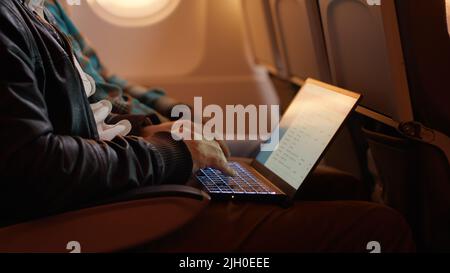 Müde Angestellte, die mit dem Flugzeug unterwegs sind und am Laptop arbeiten, während des Sonnenuntergangs mit dem Computer auf dem Flug. Geschäftsreise im Flugzeug, Flug im Urlaub in der Economy Class mit Menschen. Stockfoto