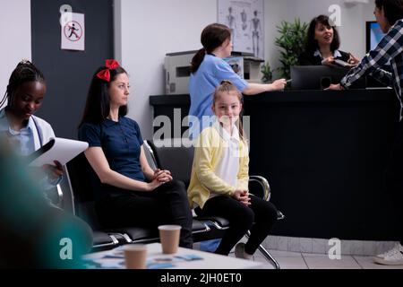 Portrait eines lächelnden jungen Mädchens mit besorgt nachdenklichen Mutter, die in einer geschäftigen Klinikrezeption mit verschiedenen Menschen sitzt. Elternteil mit Kind an der privaten Krankenhausrezeption wartet auf einen Kinderarzt. Stockfoto