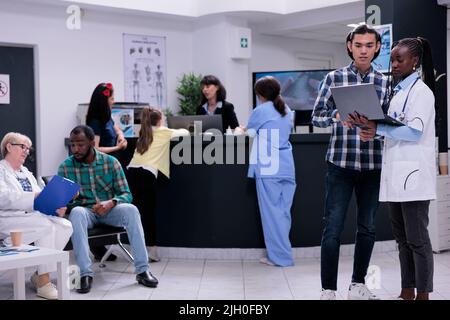 Medic hält einen Laptop mit Laborergebnissen und spricht mit einem asiatischen Patienten, während ein älterer Arzt den Patienten in eine Privatklinik einführt. Facharzt, der mit einem jungen Mann über einen Termin sprach. Stockfoto