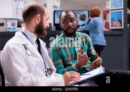 Hausarzt im Wartezimmer, der mit einem männlichen Patienten spricht und einen medizinischen Kontrollbericht zur Diagnose und Behandlung der Krankheit verwendet. Arzt und Mann, die Gesundheitsberatung durchführen. Stockfoto