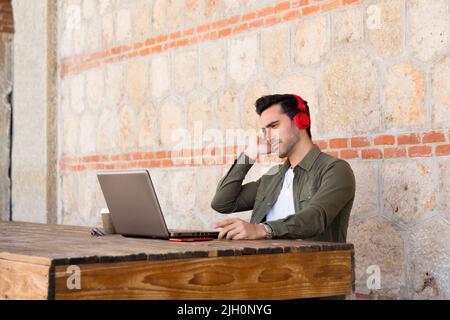 Kaukasischer Mann mit ernsthafterischem Ausdruck, der mit einem Laptop arbeitet, während er Kopfhörer auf einer Terrasse verwendet Stockfoto