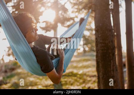 Hübscher junger Mann in einer Brille, der in der Hängematte liegt und sich beim Kaffee entspannt. Waldreisen. Stockfoto