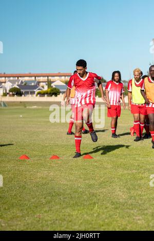 Multirassische männliche Athleten im roten Trikot, die auf dem Spielplatz gegen den klaren Himmel um Scheibenkegel springen Stockfoto