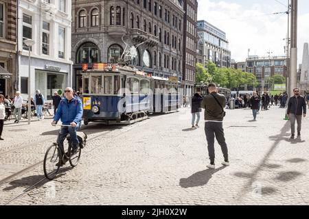 Ein Mann fährt an einer alten Straßenbahn vorbei, die auf einem belebten Dam-Platz in Amsterdam, Niederlande, geparkt ist. Stockfoto