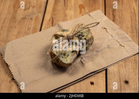 Schimmeliges Brot vor einem hölzernen Hintergrund. Zwei Scheiben Weizenbrot mit Pilzschimmel bedeckt. Die Scheiben sind mit Garn gebunden. Brauner Papierbeutel. Verdorben für Stockfoto