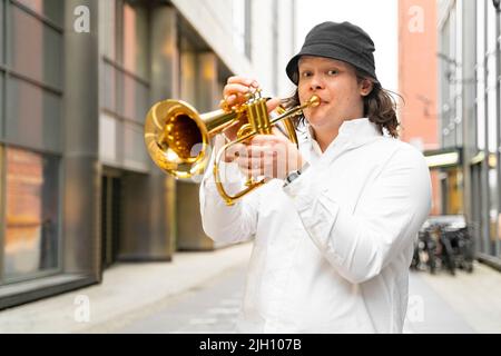 Junger kaukasischer blonder Mann mit langen Haaren, gut gekleidet, spielt Trompete im Freien Stockfoto
