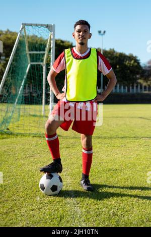 Porträt eines jungen männlichen Spielers mit einem Akimbo der Arme und einem Bein auf dem Ball über dem Grasfeld auf dem Spielplatz Stockfoto