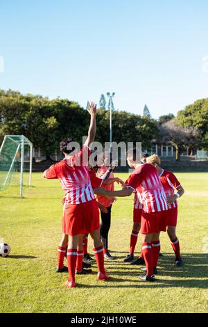 Multirassische männliche Spieler in Uniformen heben die Hände vor dem Spiel gegen den klaren Himmel auf dem Spielplatz Stockfoto