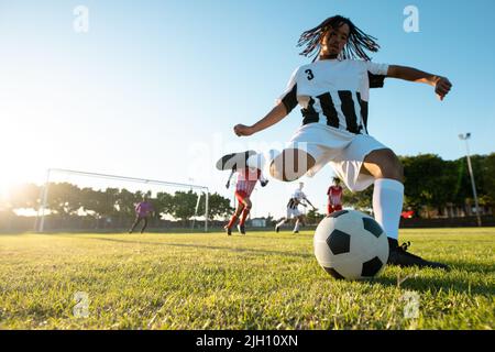 Blick aus der unteren Perspektive auf einen Spieler mit mehreren Rassen, der beim Fußballspiel gegen den klaren Himmel den Ball tritt Stockfoto