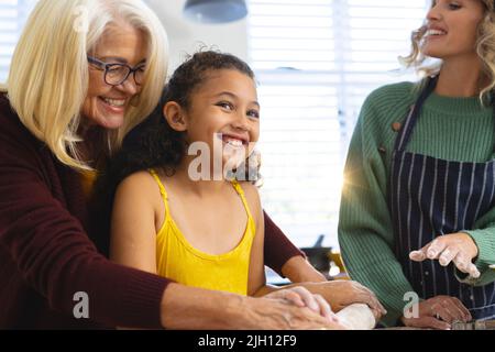 Glückliche Frau, die Großmutter ansieht, die Enkelin beim Teigrollen auf dem Tisch in der Küche unterstützt Stockfoto