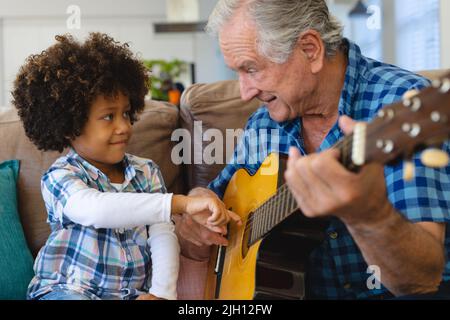 Multirassischer Großvater hält Enkels Hand und lehrt ihn, Gitarre auf dem Sofa zu Hause zu spielen Stockfoto