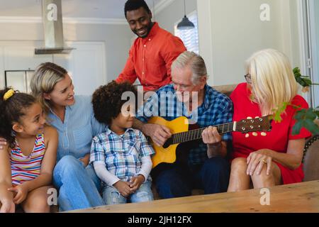 Glückliche multirassische Familie mit mehreren Generationen, die einen älteren Mann beim Gitarrenspielen ansieht, während er auf dem Sofa sitzt Stockfoto