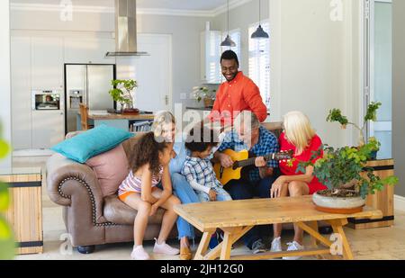 Multirassischer älterer Mann, der Gitarre spielt, während er mit einer glücklichen Familie aus mehreren Generationen auf dem Sofa sitzt Stockfoto