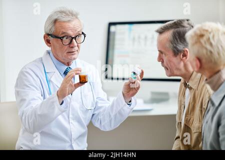 Selbstbewusster, gutaussehender Arzt im Alter, der in einem Labormantel und einer Brille zwei Pillenflaschen hält und den Patienten den Unterschied zwischen den Medikamenten erklärt Stockfoto