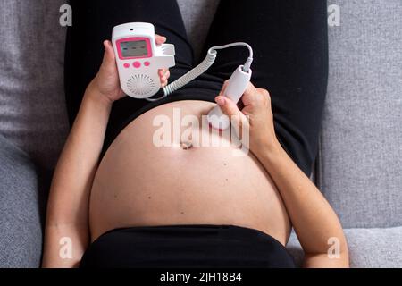 Eine Schwangerin mit einem fetalen doppler hört dem Baby zu Hause auf dem Sofa zu Stockfoto