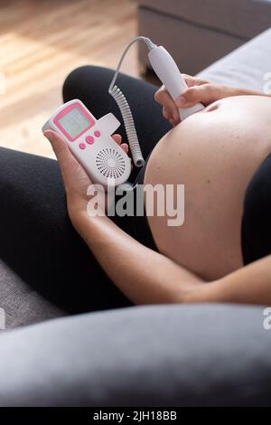 Eine Schwangerin mit einem fetalen doppler hört dem Baby zu Hause auf dem Sofa zu Stockfoto