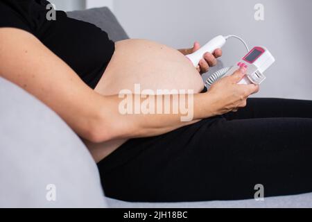 Eine Schwangerin mit einem fetalen doppler hört dem Baby zu Hause auf dem Sofa zu Stockfoto