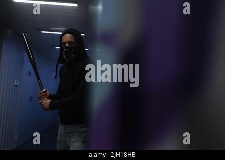 afroamerikanischer Hooligan mit Maske im Gesicht, die den Baseballschläger in der Nähe einer verschwommenen Wand in der Garage hält Stockfoto
