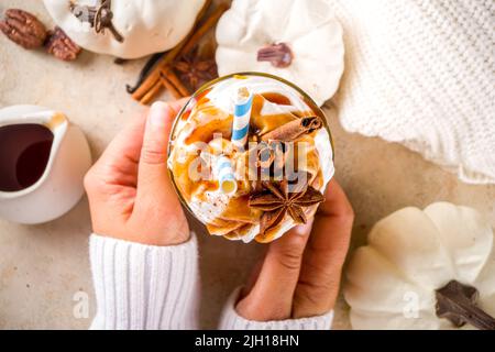 Becher mit Kürbisgewürz Latte oder Milchshake in Frauenhand, traditionelles herbstliches Heißgetränk mit Anisgeschmack, Zimt, Karamell-Belag, auf warmem, farbigem Hintergrund Stockfoto