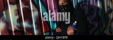 afroamerikanischer Vandalismus mit abgedecktem Gesicht, das Sprühfarbe in der Nähe von Graffiti auf der städtischen Straße hält, Banner Stockfoto