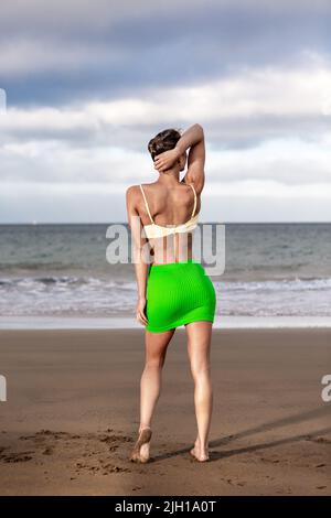 Rückansicht einer anonymen Frau in Rock und BH, die den Kopf berührt und auf das winkende Meer und den wolkigen Himmel blickt, während sie Zeit am Sandstrand verbringt Stockfoto