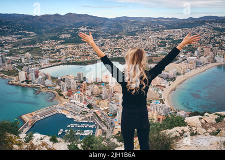 Frau auf dem Gipfel von Penon de Ifach hob ihre Hände und genießt das malerische Stadtbild von oben, Rückansicht. Tourismus, Wahrzeichen, Freiheitskonzept. Calpe, Spanien Stockfoto