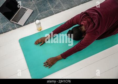 Junge schwarze afrikanische Frau macht Yoga-Übungen online. Stretching zu Hause auf der Matte vor dem Laptop-Computer, direkt von oben aufgenommen. Stockfoto