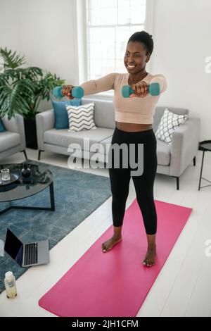 Schwarze afrikanische Frau beim Training nach Online-Fitness-Training auf dem Laptop. Sitzen Sie auf einer Yogamatte und halten Sie die Hanteln im Komfort eines modernen Wohnzimmers Stockfoto