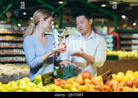 Im Supermarkt streiten sich ein multirassisches Paar, ein asiatischer Ehemann und eine asiatische Frau, die Obst und Essen auswählen, und können sich nicht auf die Auswahl von Äpfeln einigen Stockfoto