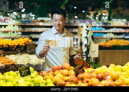 Junger asiatischer Mann wählt und pflückt im Öko-Beutel Apfelfrüchte oder Gemüse im Supermarkt. Männlicher Kunde, der einen Lebensmittelladen in der Nähe der Theke steht, kauft und wirft ein wiederverwendbares Paket auf dem Markt ein Stockfoto