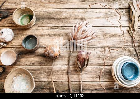 Flaches Set aus handgefertigtem, modernem Keramik-Geschirr. Draufsicht auf ungewöhnliche kreative Schalen mit seltenen trockenen Blumen auf altem Holzhintergrund. Beige und blauer EM Stockfoto