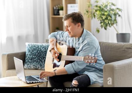 Junger Mann mit Laptop, der zu Hause Gitarre spielt Stockfoto