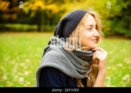 Hübsche lächelnde Frau mit Strickmütze und Schal, die in der Herbstsaison im Stadtpark nachdenklich im Freien schaut Stockfoto