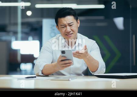 Schockiert und verärgert junger Mann asiatische Geschäftsmann Spiele auf Handy für Geld bei der Arbeit, verloren, erhielt schlechte Nachrichten durch Nachricht. Traurig am Schreibtisch in einem modernen Büro sitzen. Stockfoto