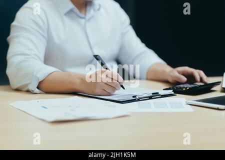 Nahaufnahme. Die Hände eines jungen Mannes ein Buchhalter in einem weißen Hemd, der an einem Schreibtisch mit Dokumenten, einem Taschenrechner, arbeitet, schreibt. Sitzen in einem modernen Büro Stockfoto