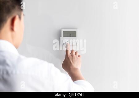 Nahaufnahme. Die Hand eines jungen Mannes in einem weißen Hemd schaltet die an der Wand hängenden Bedienknöpfe der Klimaanlage ein. Auf der linken Seite stehend Stockfoto