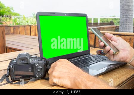 Nahaufnahme eines freiberuflichen Fotografen, der mit einem Smartphone, einem Laptop und einer leeren Spiegelreflexkamera mit grünem Bildschirm auf einem Holztisch im Freien arbeitet. Stockfoto