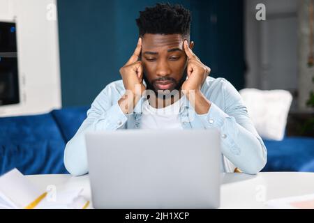 Müder, männlicher Büroangestellter, der die Hände am Kopf hält, während er auf den Laptop-Bildschirm schaut, fühlt sich langweilig und wütend. Gelangweilter multirassischer Mann in schickem Casual-Hemd, der am Schreibtisch sitzt Stockfoto