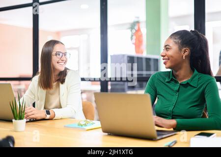 Zwei weibliche Kollegen unterhalten sich in einem modernen Büro mit offenem Raum freundlich und lächelnd, nebeneinander sitzend Stockfoto