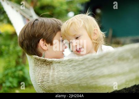 Glücklicher Kleinkind Junge und seine Schwester im Teenageralter entspannen in der Hängematte an schönen Sommertag. Nette Kinder, die Spaß im Sommerpark haben. Familienurlaub im Summe Stockfoto