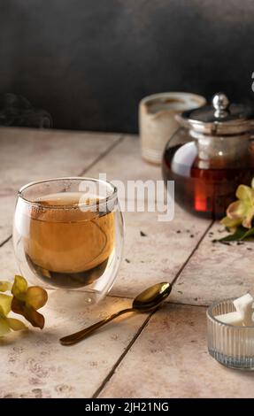 Tee aus Thermalglas mit Dampf auf beigefarbenem Fliesenhintergrund in Komposition mit Blumen, Zucker und Teekannen. Vertikale Ausrichtung. Textbereich Stockfoto