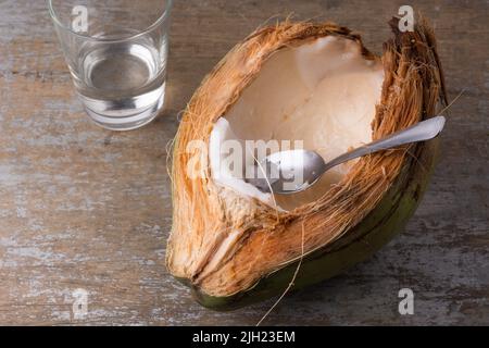 Essen von jungem Kokosfleisch direkt aus der Schale mit einem Löffel, weißes Fleisch von gesunden tropischen Früchten auf einer Holztischplatte, Platz für Text Stockfoto