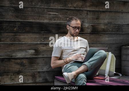 Fokussierter junger Mann, der eine Brille mit einem Laptop trägt, auf der Tastatur tippt, freiberuflicher Mitarbeiter, der online arbeitet Stockfoto