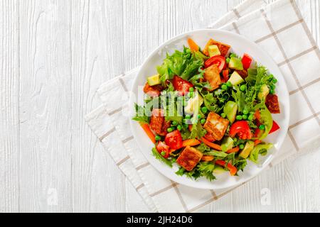 Tofu-Salat mit Avocado, knackigen Salatblättern, grünen Erbsen, Tomaten, gelber Paprika auf weißem Teller auf weißem Tisch, horizontale Ansicht von oben, f Stockfoto