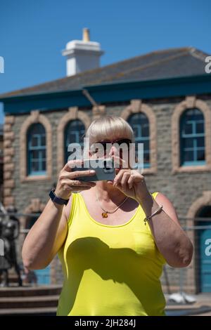 Reife Touristenfahrerin, die im Sommer im Urlaub eine Sonnenbrille und ein gelbes Oberteil trägt und Fotos auf einem Mobiltelefon fotografiert. Stockfoto
