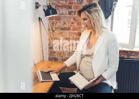 Schwanger, aber immer noch aktiv bei der Arbeit. Eine blonde kaukasische Frau mittleren Alters in einem weißen Blazer sitzt an ihrem Schreibtisch, arbeitet an einem Laptop und hält einen Taschenbuchkalender. Hochwertige Fotos Stockfoto