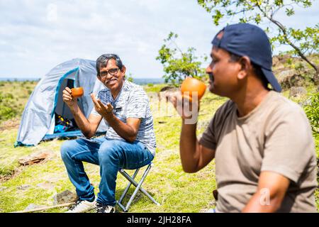 Wanderer mittleren Alters bei einem Tee, während sie sich in der Nähe des Campingzeltes am Ziel während der Wochenendferien unterhalten - Konzept der Erfrischung, Freundschaft Stockfoto